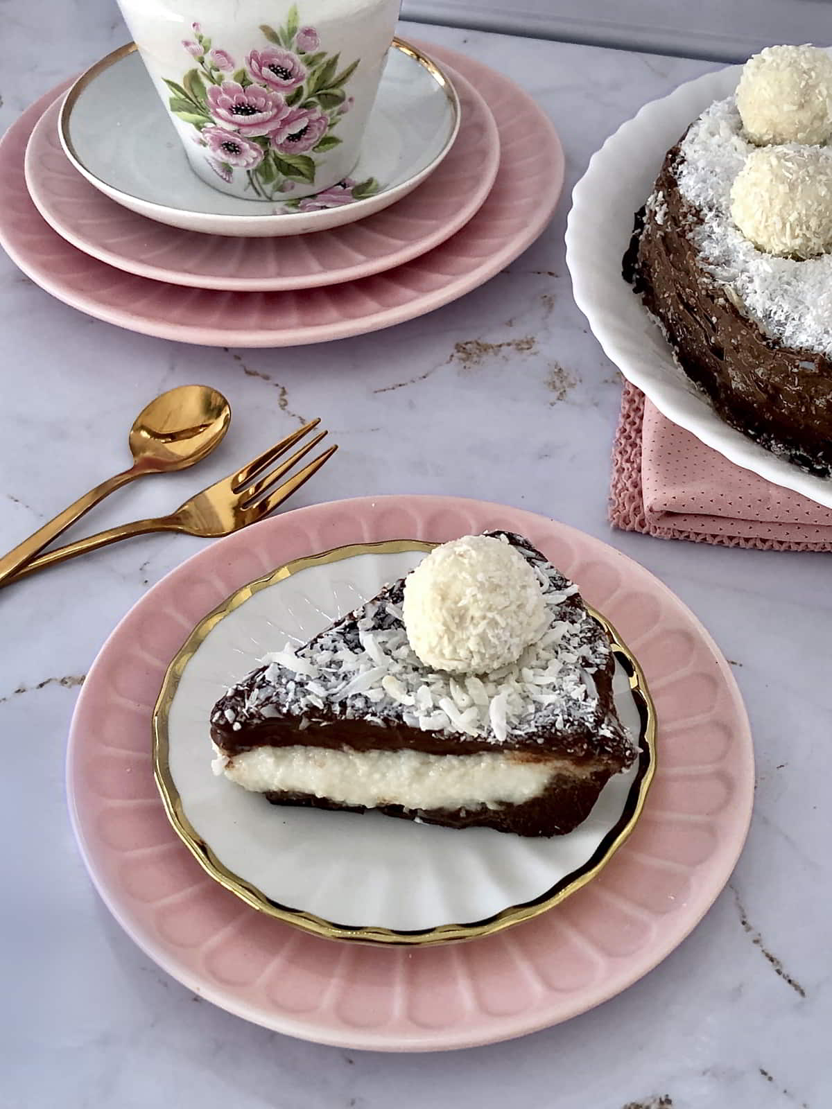 Slice of coconut cream pie with chocolate ganache and shredded coconut on a pink plate beside a floral teacup — a calm and elegant dessert moment.